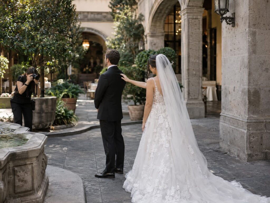 Novia tocando la espalda del novio de espaldas en un patio colonial justo antes del First Look mientras la fotógrafa captura el momento